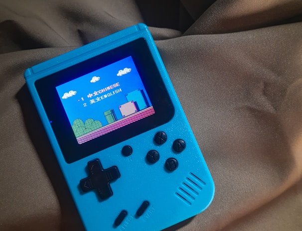 A handheld gaming device with a bright blue casing rests on a soft, dark fabric background. The screen displays a pixelated video game scene with options to select either Chinese or English language. The layout includes a classic D-pad and buttons, reminiscent of retro gaming consoles.